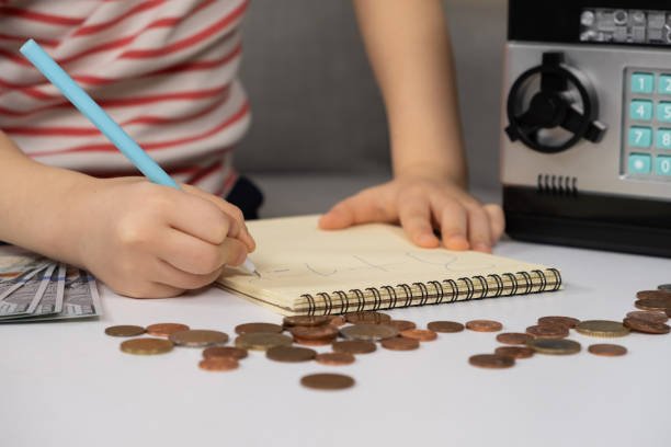 A child writing a savings plan in a notebook beside coins and a small safe